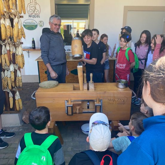 Ein älterer Mann demonstriert eine Holzpresse für eine Gruppe von Kindern und Erwachsenen in einem Werkstattumfeld. Ein Tisch mit einem Fass und einem hölzernen Objekt ist im Vordergrund. Mais hängt an der Wand.