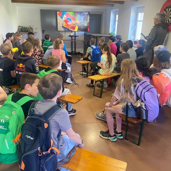 Schüler sitzen in einem Klassenzimmer mit Holzbänken und Rucksäcken und sehen einem großen Bildschirm, der ein farbenfrohes Bild zeigt. Ein Erwachsener steht in der Nähe des Bildschirms und spricht die Gruppe an.
