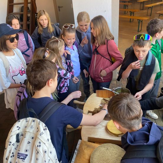 Eine Gruppe von Kindern versammelt sich um einen Tisch, wahrscheinlich in einer Schule. Sie scheinen Essen zuzubereiten. Einige von ihnen tragen Rucksäcke und Sonnenbrillen. Der Tisch hat ein Schneidebrett und einen Topf. Hinter ihnen befindet sich eine Tür und eine Leiter.
