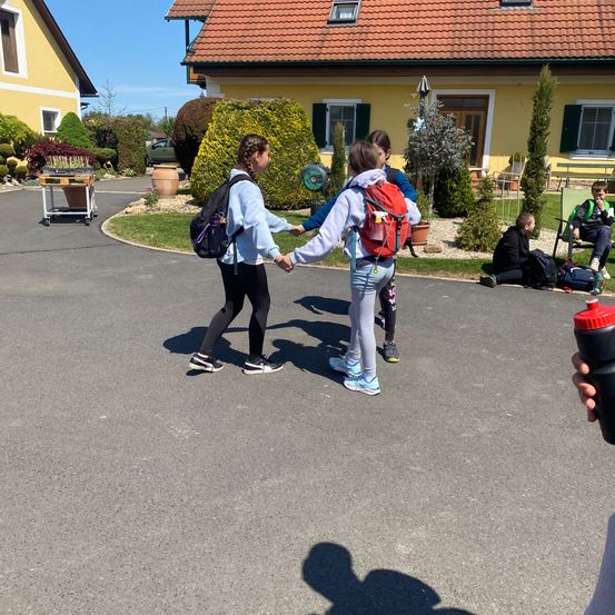 Drei Menschen, wahrscheinlich Freunde, halten sich an den Händen, während sie auf der Straße gehen. Im Hintergrund befindet sich ein Haus mit rotem Dach.
