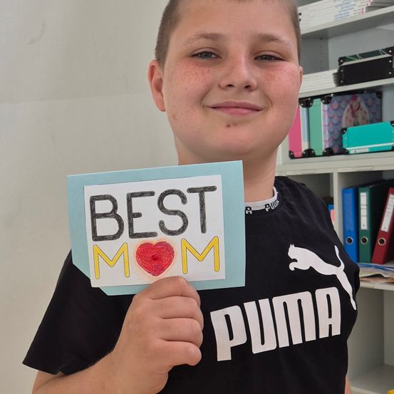 Ein junger Junge hält eine selbstgemachte Karte mit der Aufschrift 'Best Mom' mit einem roten Herzen auf dem 'o'. Er trägt ein schwarzes Puma-Shirt.