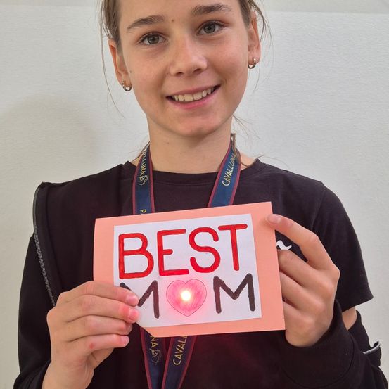 Ein Mädchen hält eine rosa Karte, auf der 'Beste Mama' mit einem Herz und einem Licht darauf steht. Sie lächelt und posiert für ein Foto.