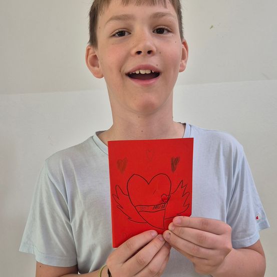 Ein Junge hält einen roten Valentinstags-Karte mit einem Herz und lächelt für die Kamera.