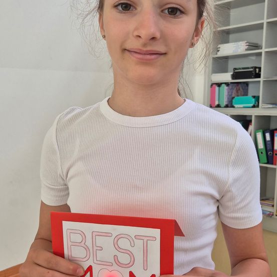 Eine Frau in einem weißen T-Shirt lächelt, während sie eine rote Karte mit der Aufschrift BEST MOM in der Hand hält. Dahinter steht ein weißer Regal mit Büchern und Ordnern.