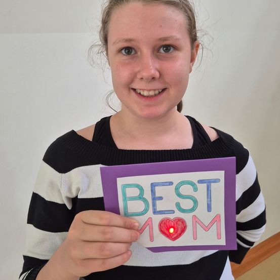 Ein strahlendes Mädchen hält einen Karton mit 'BESTE MUTTER' in großen Buchstaben, einem Herzsymbol und einem leuchtenden roten Licht im Herzen. Sie trägt einen schwarz-weiß gestreiften Pullover.