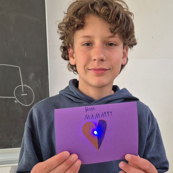 Ein junger Junge hält eine lila Karte mit einem Herz und einem Licht darauf hoch. Er lächelt und trägt eine blaue Kapuzenjacke. Hinter ihm befindet sich eine Tafel mit einigen Zeichnungen.