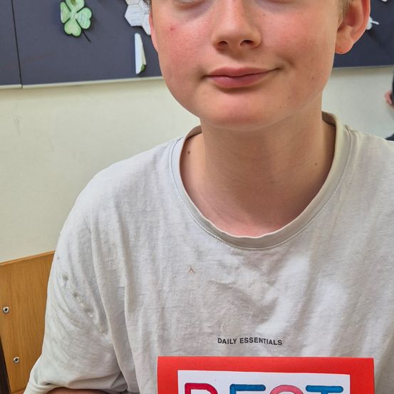 Ein junger Junge trägt ein weißes T-Shirt mit Daily Essentials und hält ein rotes Best-Poster vor seiner Brust.