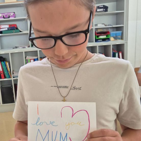 Eine Person hält eine handgemachte Karte mit der Botschaft 'Ich liebe dich MUM'. Die Person trägt eine Brille und ein weißes T-Shirt. Dahinter steht ein weißer Regal mit verschiedenen Gegenständen.