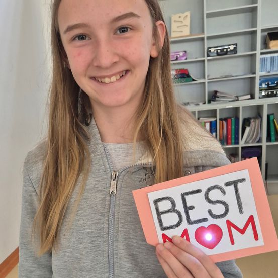 Ein Mädchen lächelt und hält ein Schild mit den Worten BESTE MUTTER, das von einem roten Licht beleuchtet wird. Sie trägt eine graue Kapuzenjacke. Im Hintergrund befindet sich ein weißer Bücherregal mit Büchern und Ordnern.