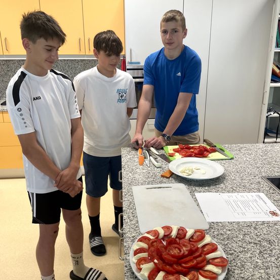 Drei Jungen bereiten Essen in einer Küche zu. Einer steht und beobachtet, während die anderen beiden Tomaten auf einem Tresen schneiden. Ein Teller mit geschnittenen Tomaten ist in der Nähe.
