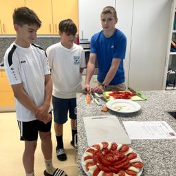 Drei Jungen bereiten Essen in einer Küche zu. Einer steht und beobachtet, während die anderen beiden Tomaten auf einem Tresen schneiden. Ein Teller mit geschnittenen Tomaten ist in der Nähe.