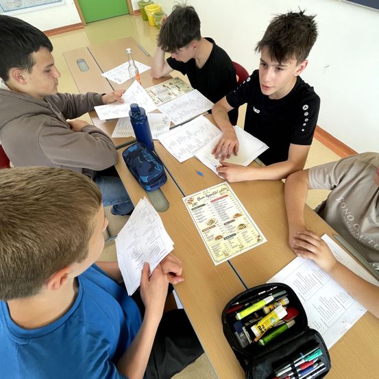 Fünf Jungen sitzen um einen Tisch herum, jeder hält Papiere und eine Speisekarte. Sie scheinen an einer Schulaktivität beteiligt zu sein. Ein Junge hat eine blaue Flasche auf dem Tisch.