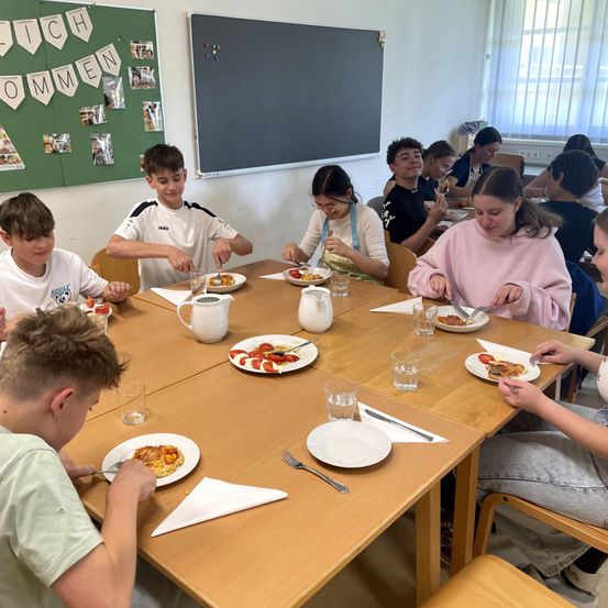 Eine Gruppe von Schülern sitzt an einem Tisch in einem Klassenzimmer und isst Pizza. Der Tisch ist mit Tellern, Gläsern und Besteck gedeckt. Das Klassenzimmer hat eine grüne Pinnwand mit verschiedenen Bildern und eine Tafel.