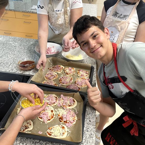 Eine Gruppe von Menschen in einer Schürze bereitet Mini-Pizzas mit Schinken und Mais auf einer Küchentheke zu.