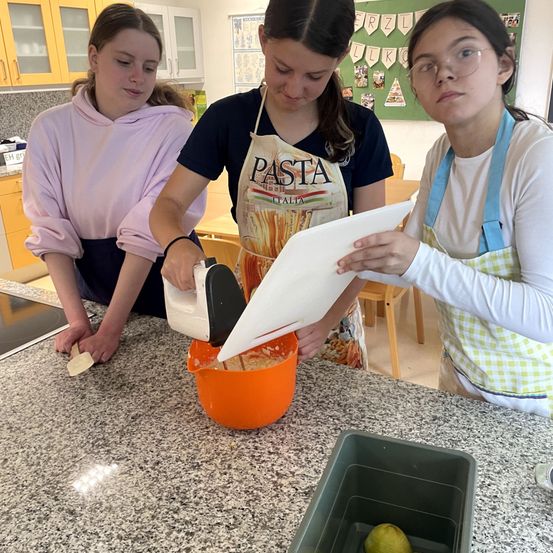 Drei Mädchen sind in einer Küche und bereiten Essen zu. Eine hält einen Topf, eine andere ein Rührgerät und die dritte hält ein Papier. Sie tragen Schürzen und haben Küchenutensilien in der Nähe.