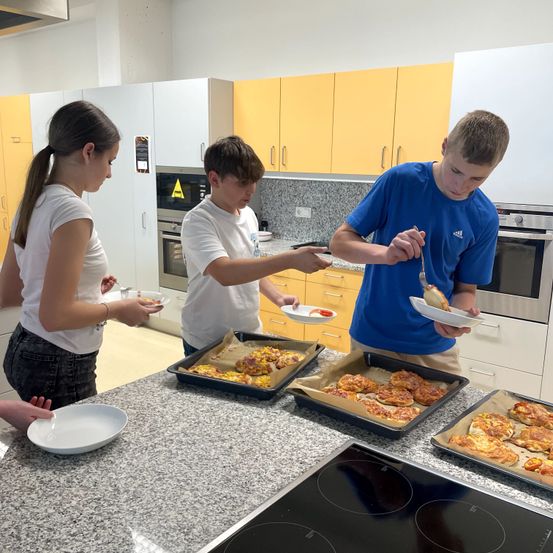 Drei Teenager sind in einer Küche. Einer hält einen Teller mit einem Essensgegenstand, ein anderer ist dabei, mit einer Gabel zu essen, und der dritte hält eine Schüssel. Die Küche hat gelbe Schränke, einen Herd und Tabletts mit Essen.