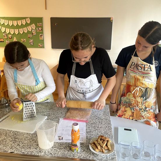 Drei Mädchen sind in einer Küche und beschäftigen sich mit der Essenszubereitung. Eines der Mädchen schneidet Käse, ein anderes rollt Teig aus, und das dritte schaut zu. Auf dem Tisch liegen verschiedene Küchenutensilien, darunter ein Messbecher, eine Flasche, eine Schüssel mit Essen und Gläser. Ein Anschlagbrett steht hinter ihnen.