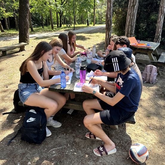 Eine Gruppe von Schülern sitzt um einen Picknicktisch in einem Park. Sie sind mit Stiften, Papieren und Wasserflaschen an einer Schreibaktivität beteiligt. Der Hintergrund zeigt Bäume und einen Weg aus Erde.