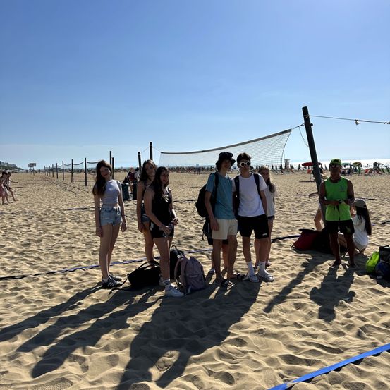 Eine Gruppe von Freunden steht auf einem sandigen Strand mit Volleyballnetzen im Hintergrund. Sie tragen lässige Sommerkleidung.