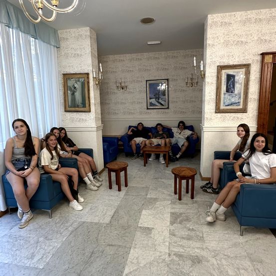 Eine Gruppe von Menschen sitzt in einem Raum mit blauen Sofas und Holzstühlen. Der Raum hat Marmorböden, weiße Wände mit Bilderrahmen und eine Kronleuchter.