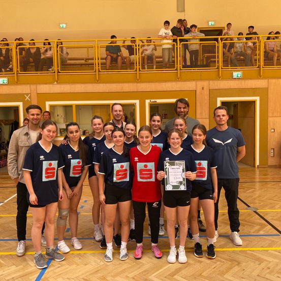 Eine Gruppe junger Athleten mit Trainern posiert für ein Foto auf einem Sportplatz. Die Athleten tragen passende Trikots und Shorts. Ein Mädchen in der Mitte hält ein Zertifikat. Zuschauer beobachten von der Galerie aus.