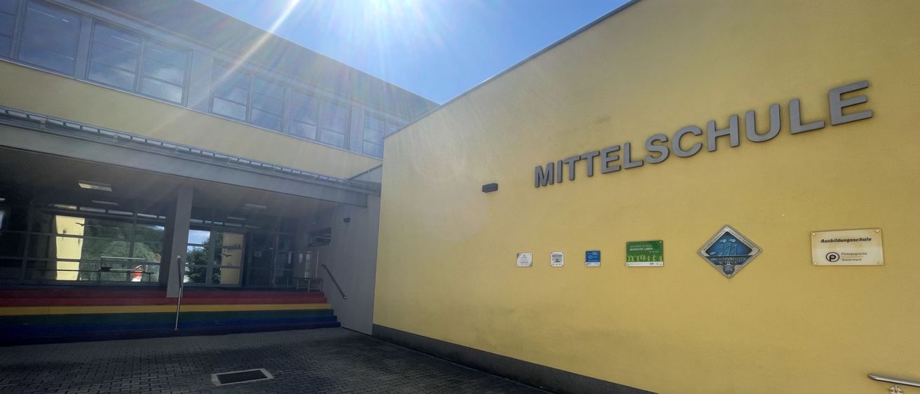 Ein hell beleuchtetes Bild eines Schulgebäudes an einem sonnigen Tag. Das Gebäude hat eine gelbe Außenfassade und ist mit 'Mittelschule' beschriftet. Eine farbenfrohe Treppe führt zum Eingang.