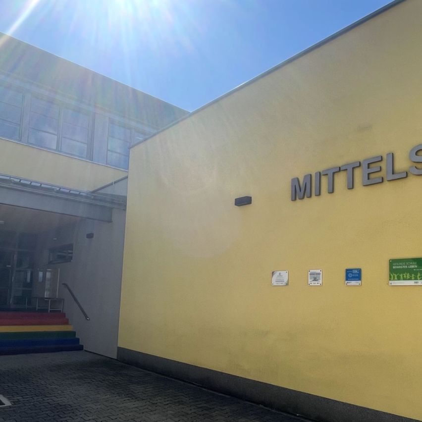 Ein hell beleuchtetes Bild eines Schulgebäudes an einem sonnigen Tag. Das Gebäude hat eine gelbe Außenfassade und ist mit 'Mittelschule' beschriftet. Eine farbenfrohe Treppe führt zum Eingang.
