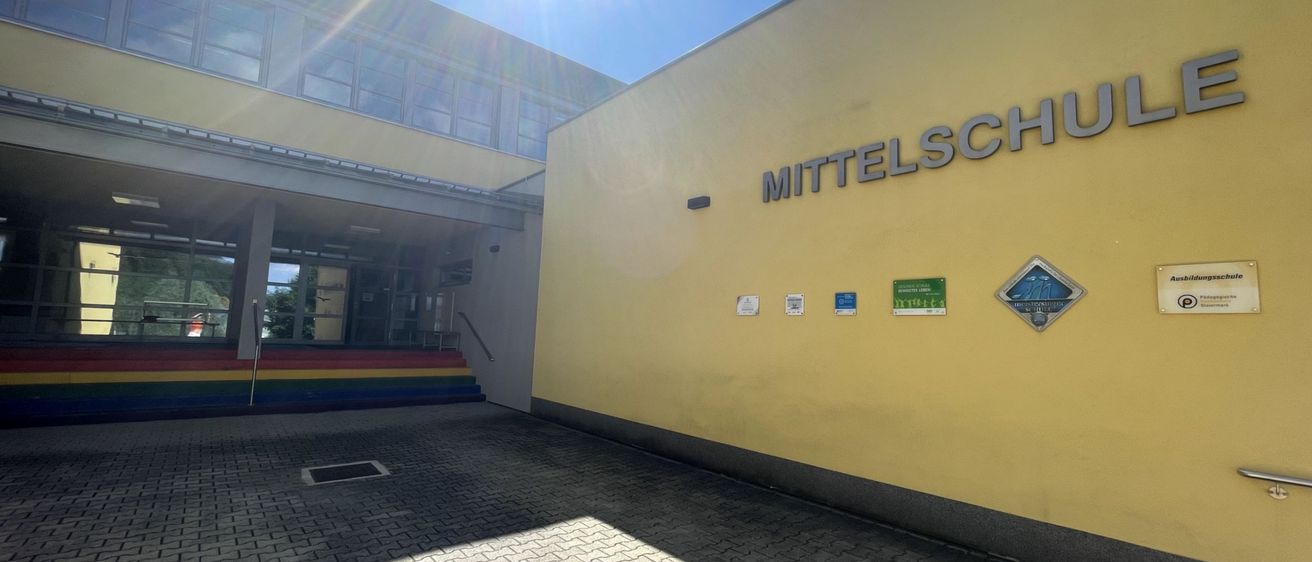 Ein hell beleuchtetes Bild eines Schulgebäudes an einem sonnigen Tag. Das Gebäude hat eine gelbe Außenfassade und ist mit 'Mittelschule' beschriftet. Eine farbenfrohe Treppe führt zum Eingang.