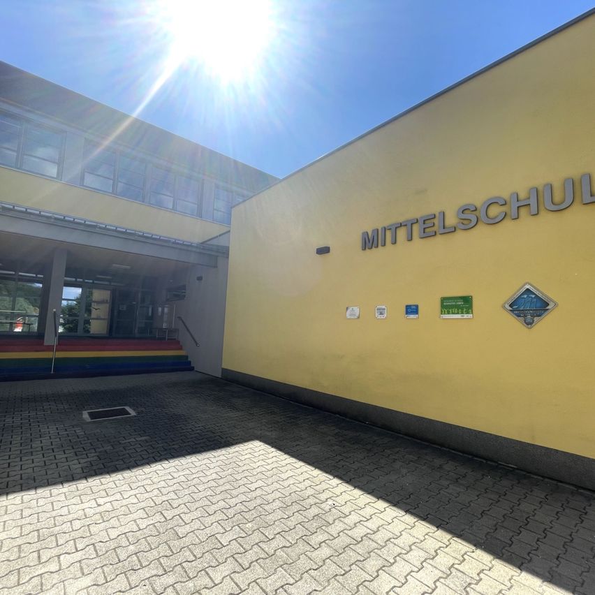 Ein hell beleuchtetes Bild eines Schulgebäudes an einem sonnigen Tag. Das Gebäude hat eine gelbe Außenfassade und ist mit 'Mittelschule' beschriftet. Eine farbenfrohe Treppe führt zum Eingang.