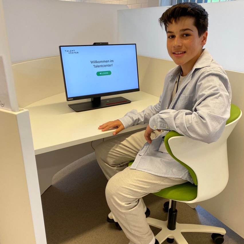 Ein junger Mann sitzt in einer Kabine mit einem Schreibtisch und lächelt in die Kamera. Ein Monitor zeigt 'Willkommen im Talentcenter!'