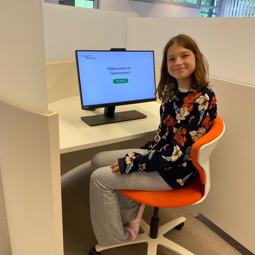 Ein junges Mädchen in einem Blumenhemd und grauen Hosen sitzt in einem Bürostuhl vor einem Computer. Der Monitor zeigt 'Willkommen im Talentcenter!' und eine grüne Login-Schaltfläche.