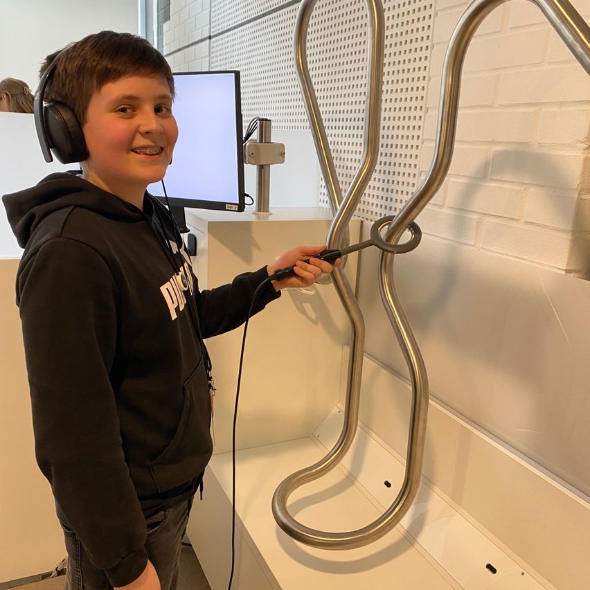 Ein junger Junge mit Kopfhörern steht in einem Raum, hält ein Werkzeug und lächelt, mit einem Monitor und einer Metallstruktur hinter ihm.