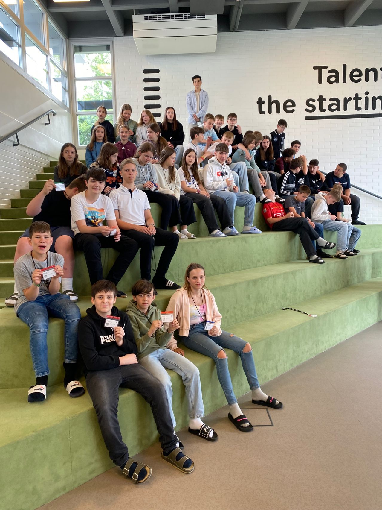 Eine Gruppe von Schülern sitzt auf grünen Bänken in einem Gebäude. An der Wand dahinter steht 'Talent the starting'. Jeder Schüler hält ein Stück Papier.