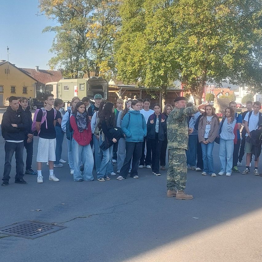Eine Gruppe von Schülern ist draußen versammelt, wobei ein Militäroffizier sie anspricht. Die Umgebung ist draußen, mit Bäumen und Gebäuden im Hintergrund.