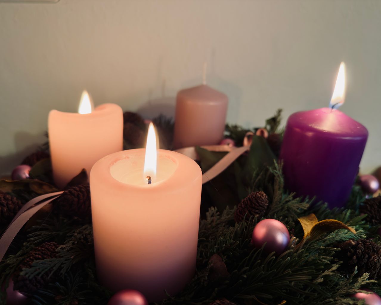 Ein Kranz aus immergrünen Pflanzen hält vier brennende Kerzen. Zwei Kerzen sind rosa, eine ist lila und eine ist rosa mit einem weißen Docht. Der Kranz hat Tannenzapfen und Stechpalmenblätter.