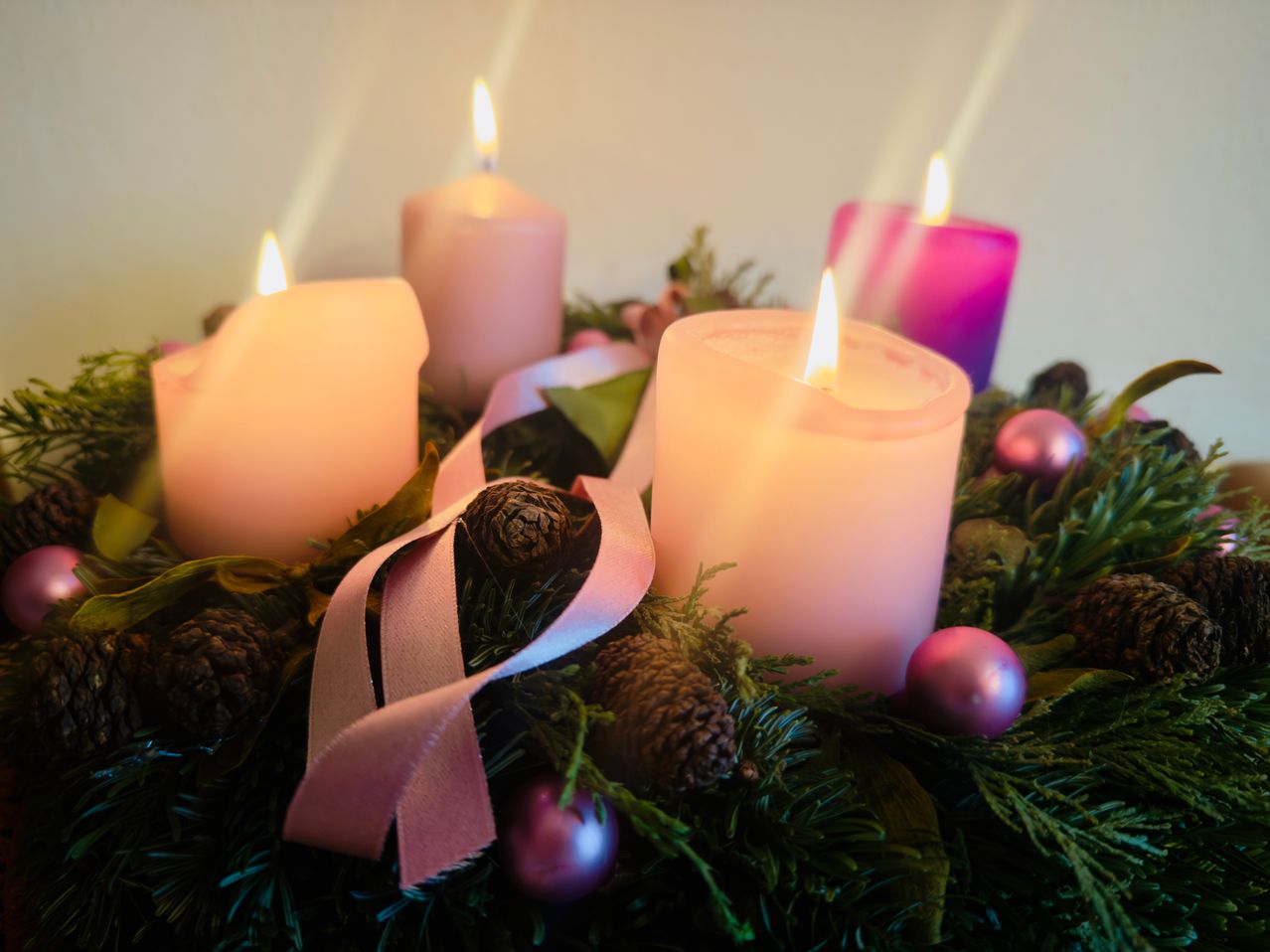 Ein Kranz aus immergrünen Pflanzen mit vier brennenden Kerzen und dekorativen Ornamenten. Ein rosafarbenes Band ist über den Kranz gelegt. Die Kerzen sind rosa und lila.