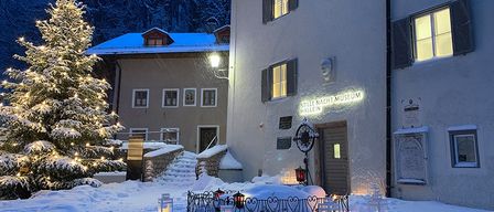 Eine verschneite Landschaft zeigt einen mit Lichtern geschmückten Weihnachtsbaum vor dem Stille Nacht Museum. Das Gebäude hat ein schneebedecktes Dach und ist mit Lampen beleuchtet.