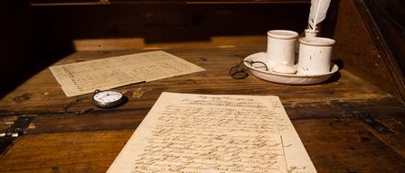 Ein alter Holzschreibtisch mit Papieren, einer Feder, Tintenpott, einer Taschenuhr und einer Brille. Dahinter befinden sich Holzschubladen.