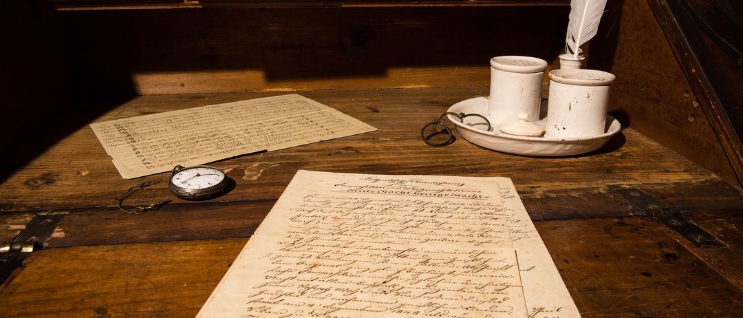 Ein alter Holzschreibtisch mit Papieren, einer Feder, Tintenpott, einer Taschenuhr und einer Brille. Dahinter befinden sich Holzschubladen.