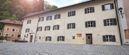 Ein großes weißes Gebäude mit mehreren Fenstern und schwarzen Fensterläden steht auf einer Kopfsteinpflasterstraße unter einem blauen Himmel.