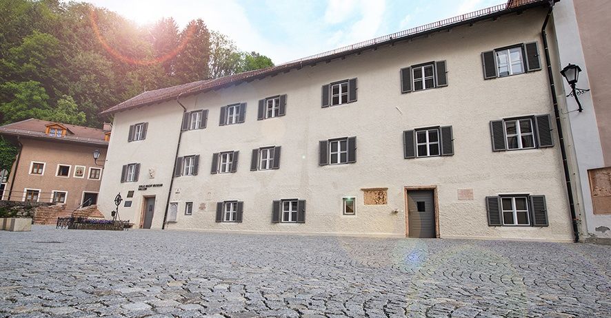 Ein großes weißes Gebäude mit mehreren Fenstern und schwarzen Fensterläden steht auf einer Kopfsteinpflasterstraße unter einem blauen Himmel.
