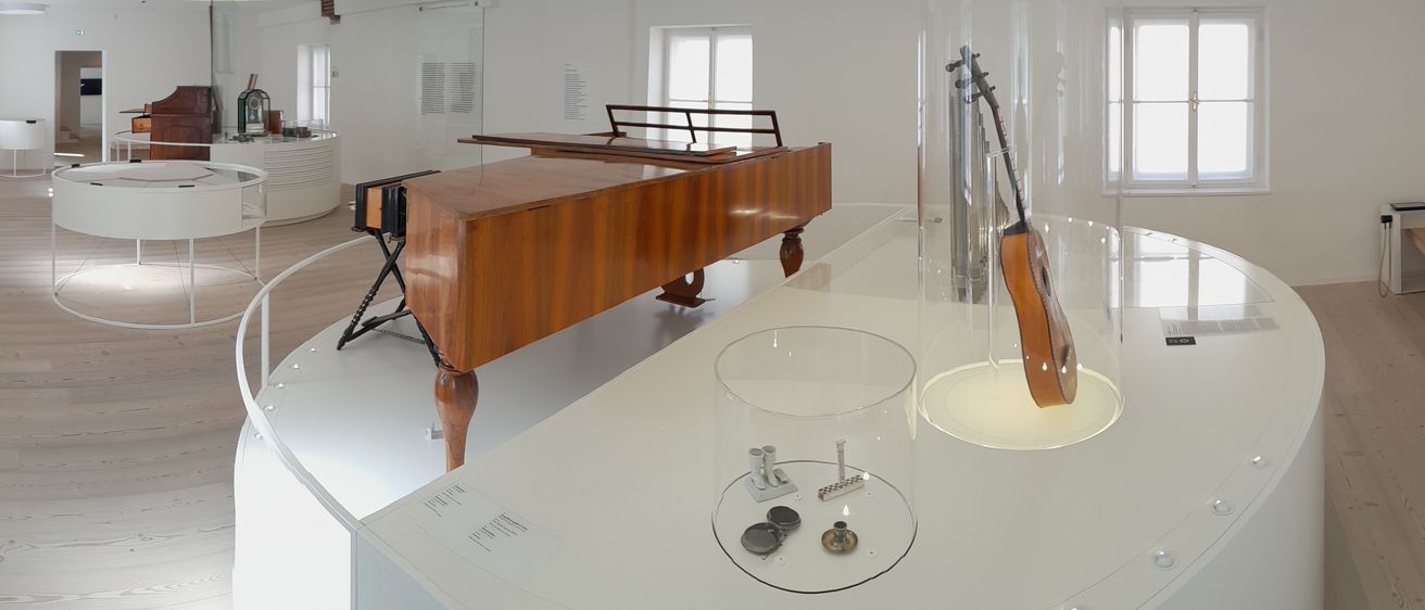 Ein modernes, minimalistisches Interieur mit einem ausgestellten Holzklavier, einem Glasvitrine und anderen Musikinstrumenten. Der Raum ist weiß mit einem Holzboden.