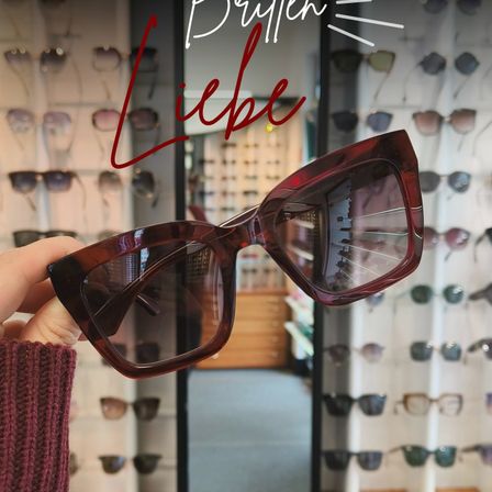 Eine Hand hält ein Paar rote Cat-Eye-Sonnenbrille, mit 'Brüllen Liebe' darüber geschrieben. Dahinter sind mehrere Sonnenbrillen an einer Wand ausgestellt.
