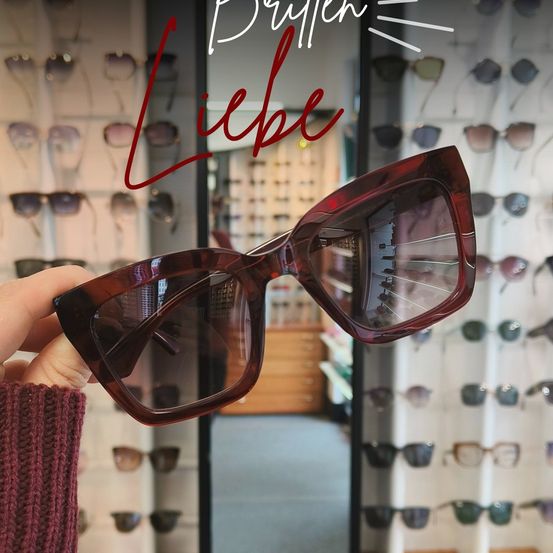 Eine Hand hält ein Paar rote Cat-Eye-Sonnenbrille, mit 'Brüllen Liebe' darüber geschrieben. Dahinter sind mehrere Sonnenbrillen an einer Wand ausgestellt.
