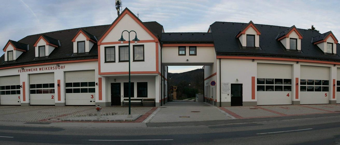 Ein zweistöckiges Gebäude mit schwarzem Dach, weißen Wänden und orangefarbenen Akzenten ist zu sehen. Der Eingang ist gewölbt, mit einer schwarzen Tür und einer Straßenlaterne. Dahinter führt eine Straße zu einer Bergkette.