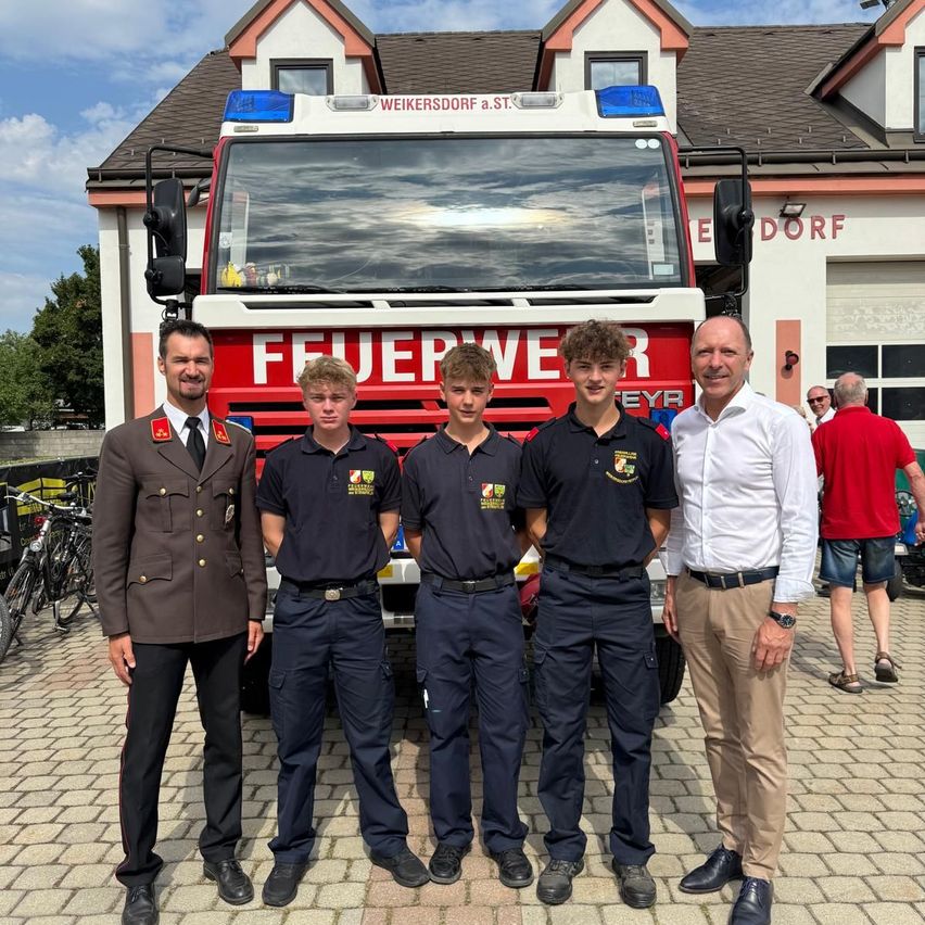 Fünf Männer stehen vor einem Feuerwehrauto. Der Mann auf der linken Seite trägt eine Militäruniform, während die anderen Feuerwehruniformen tragen. Sie lächeln für ein Foto.