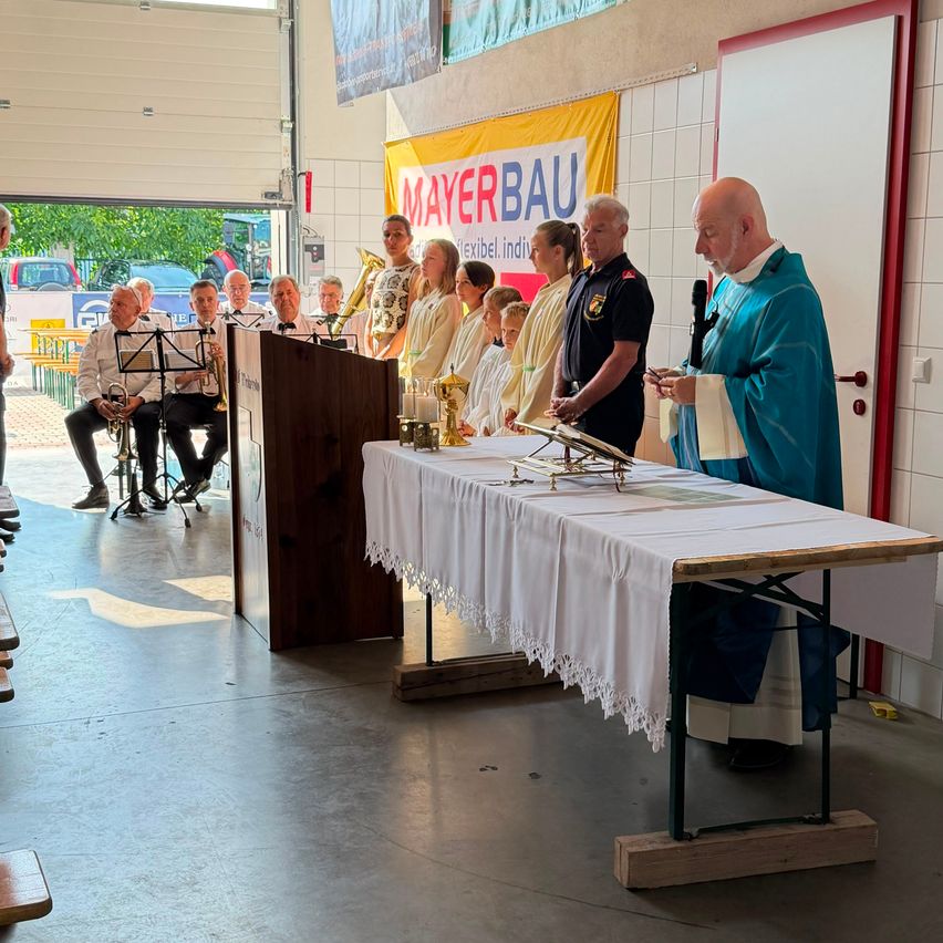Ein Priester steht mit einem Mikrofon an einem Tisch, umgeben von einer Gruppe von Menschen in einem Raum mit einem Banner, das 'Mayer Bau' liest. Einige Leute sitzen auf Stühlen mit Musikinstrumenten.