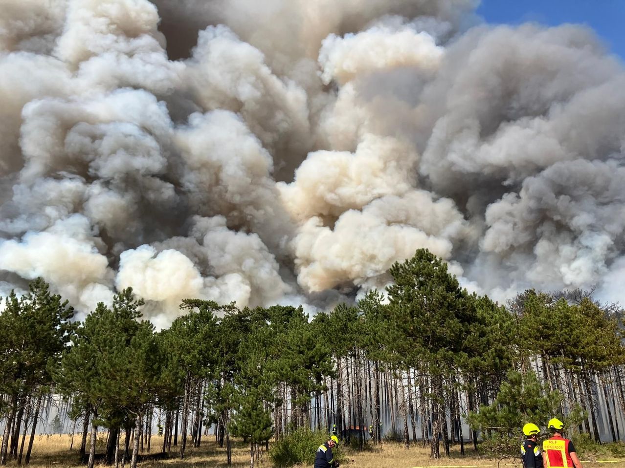 A forest fire with large smoke clouds, firefighters in yellow helmets are working to control the blaze.
