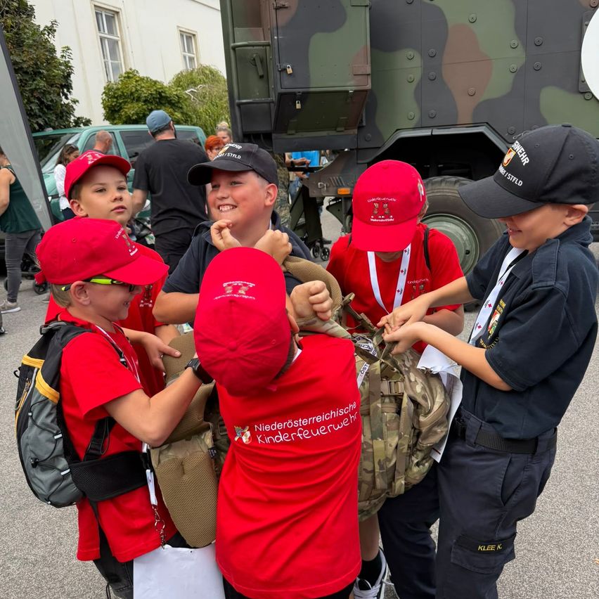 Fünf junge Jungen in roten Hemden und Mützen helfen einem Jungen in einer blauen Uniform, einen Rucksack und eine Tasche zu tragen. Sie stehen vor einem getarnten Militärfahrzeug.