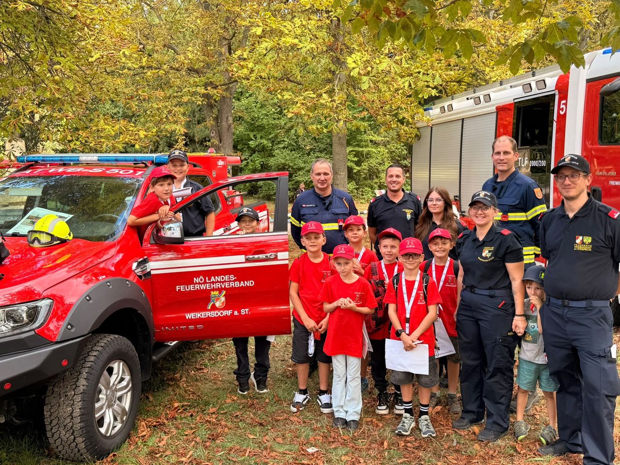 Eine Gruppe von Kindern und Erwachsenen posiert für ein Foto vor einem roten Feuerwehrauto in einem Park. Die Kinder tragen rote Mützen und Hemden, während die Erwachsenen in Uniformen sind. Das Feuerwehrauto hat den Text 'NO LANDES-FEUERWEHRVERBAND WEIKERSDORF & ST.' darauf.